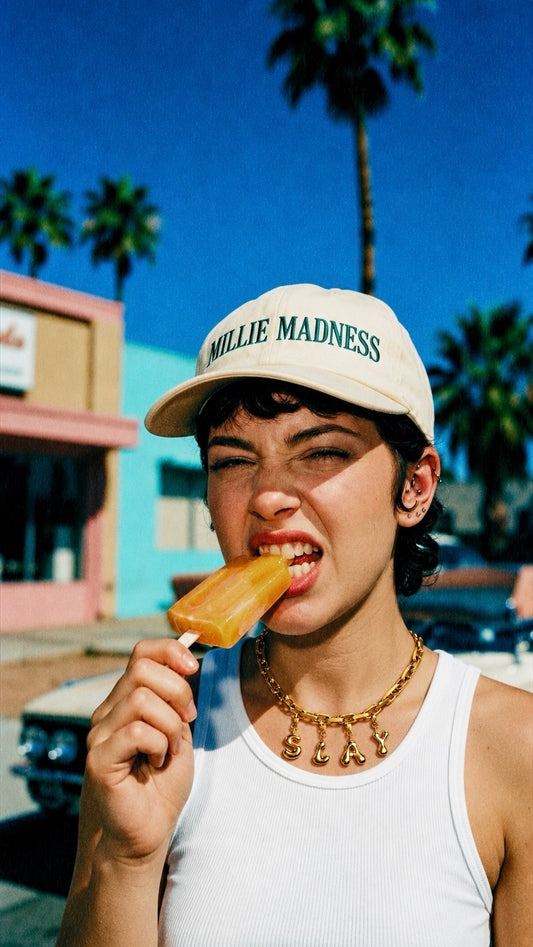 A woman outdoors wearing a cream baseball cap with green 'MILLIE MADNESS' text and a white tank top, while biting an orange popsicle. Palm trees and a bright blue sky are in the background.