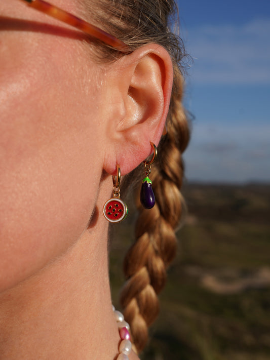 Close-up of a person's ear with earrings, watermelon earring and an aubergine earring, blurred natural background