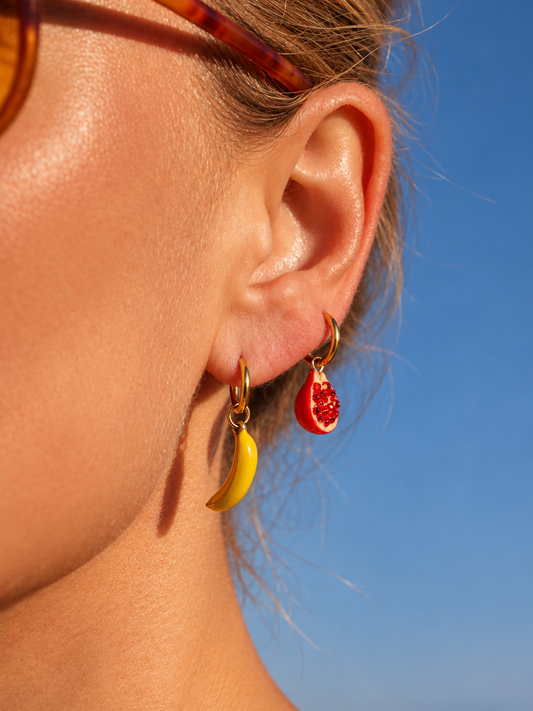A close-up of a person's ear wearing two small hoop millie madness earrings: one yellow banana-shaped earring and one red pomegranate-shaped earring, against a clear blue sky.