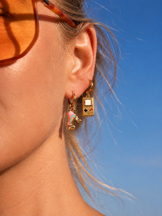 A close-up side profile of a woman wearing gold-toned dangling earrings, one shaped like a classic Game Boy and the other like a small unicorn, against a bright blue sky.