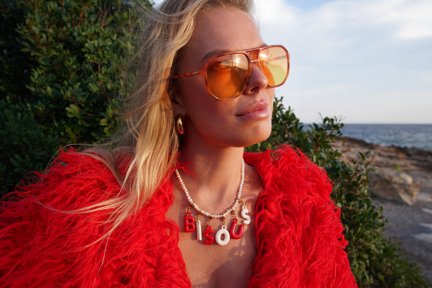Woman wearing orange sunglasses and a red jacket with 'BLESSED' necklace by the ocean.