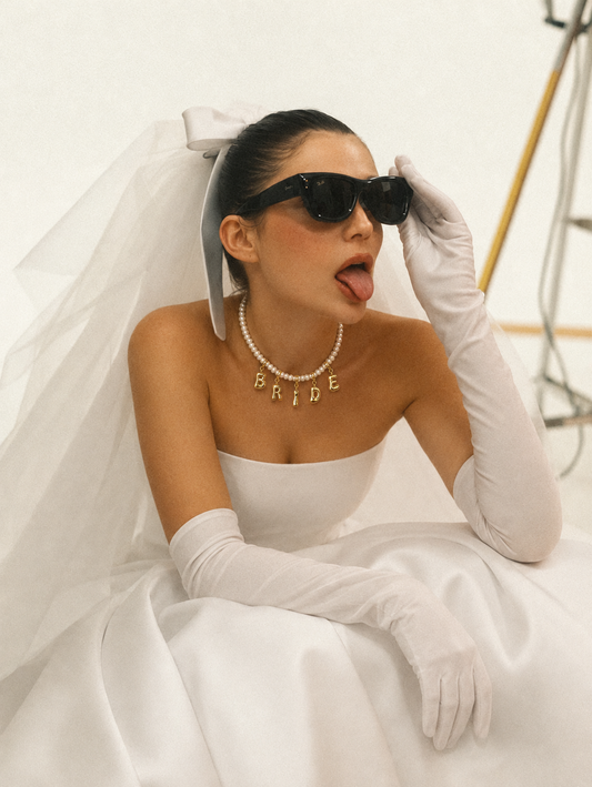 A woman in a white bridal gown with a veil, white gloves, and black sunglasses poses in a studio setting. She is wearing a pearl necklace with the word 'BRIDE' spelled out in gold letters.