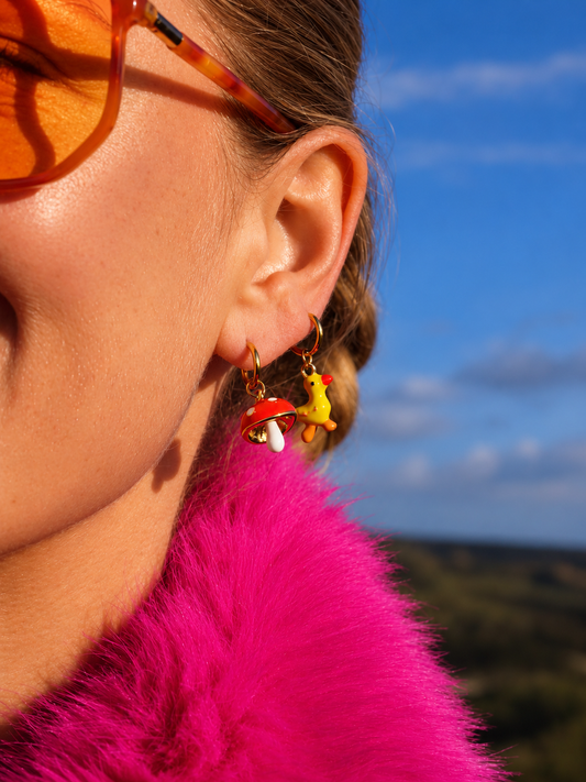 A close-up side profile of a woman outdoors wearing colorful dangling earrings with a red mushroom and a yellow chick charm, a bright pink faux-fur collar, and orange-tinted sunglasses against a blue sky background.