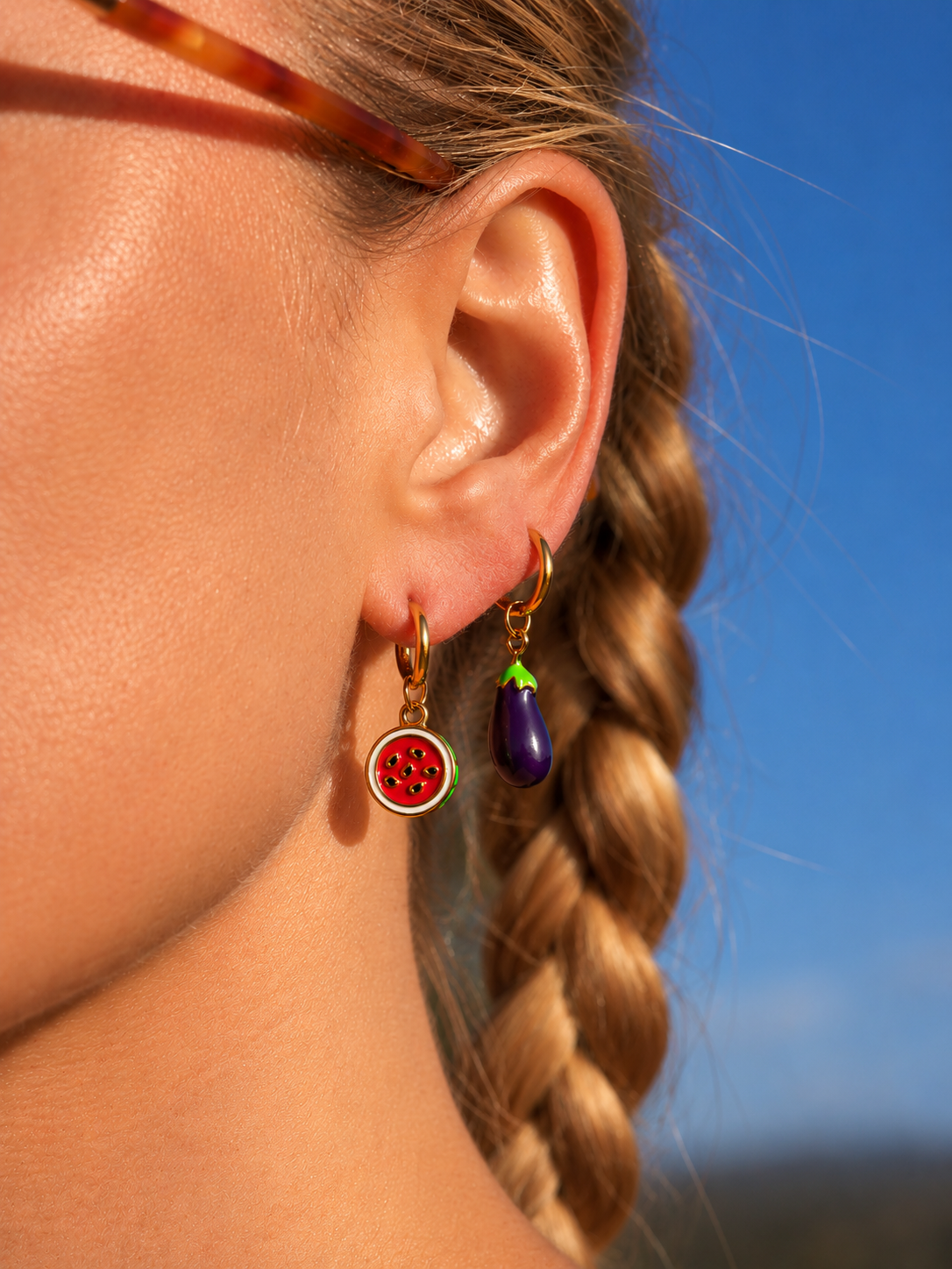 A close-up of a woman's ear wearing two colorful dangling millie madness earrings: a gold hoop with a red watermelon charm and a gold hoop with a purple eggplant charm, against a bright blue sky background.