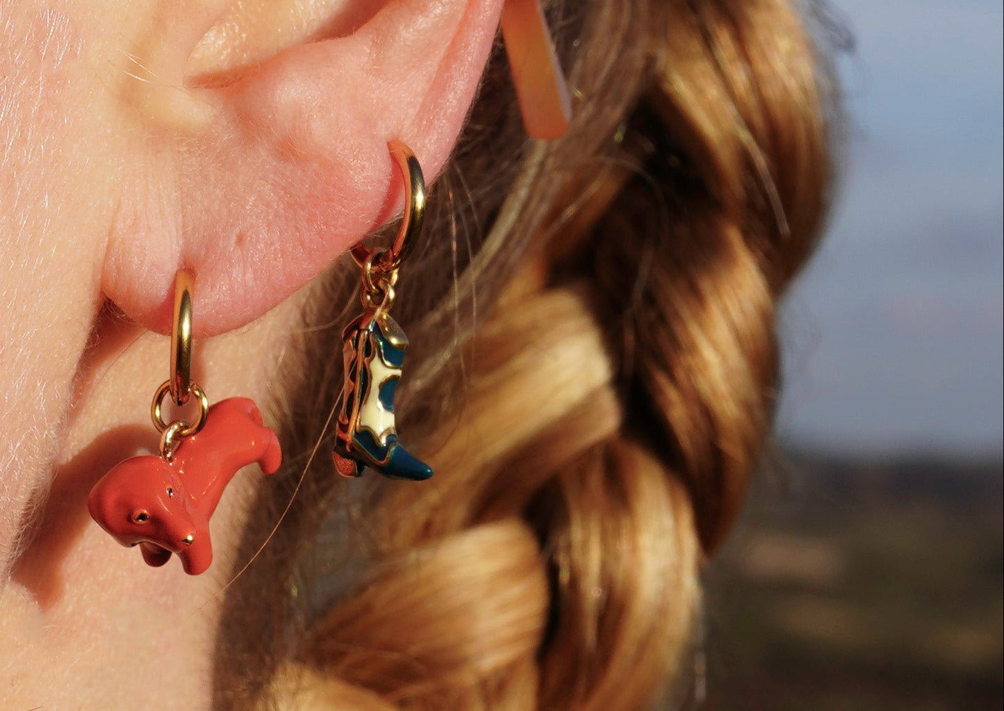 Close-up of a person's ear with earrings, wearing sunglasses, against a blurred natural background.