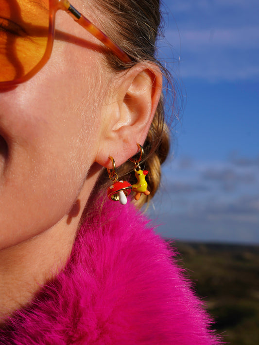 Close-up of a person wearing sunglasses and colorful earrings with a mushroom earring and a duck earring with a blurred natural background