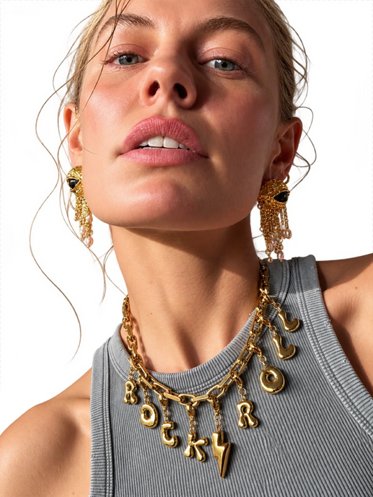A close-up of a model wearing a chunky gold millie madness necklace with dangling letter charms spelling 'ROCKY' and a lightning bolt, along with matching dangling earrings, against a clean white background.