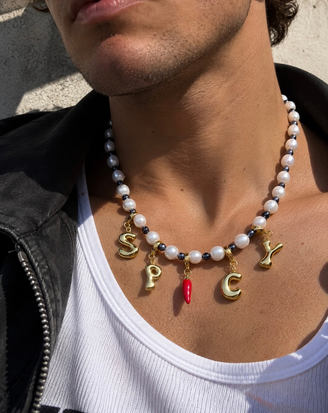 A close-up lifestyle shot of a man wearing a millie madness beaded necklace with gold letter charms spelling 'SPICY' and a small red chili pepper charm, styled with a black leather jacket and white tank top.
