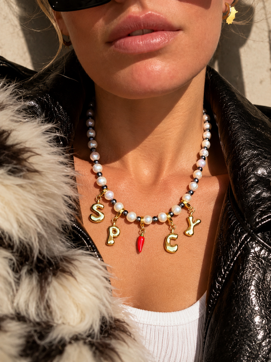 A close-up lifestyle shot of a model wearing a beaded millie madness necklace with gold letter charms spelling 'SPICY' and a small red chili pepper charm, styled with a black leather jacket and white top.