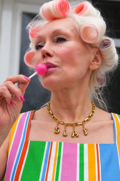 A close-up lifestyle shot of a woman with white hair in pink and orange curlers, wearing a colorful striped top and a gold chain MILLIE MADNESS necklace with the letters 'S L A Y' hanging from it. She is holding a pink lollipop.