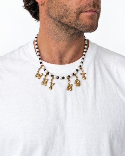 A close-up of a man wearing a beaded MILLIE MADNESS necklace with gold letter charms spelling 'WHY NOT' over a white t-shirt.