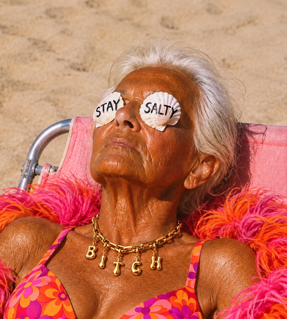 An older woman reclining on a pink beach chair on the sand, wearing shell-shaped sunglasses with the words 'STAY SALTY' and a gold necklace spelling 'BITCH'.