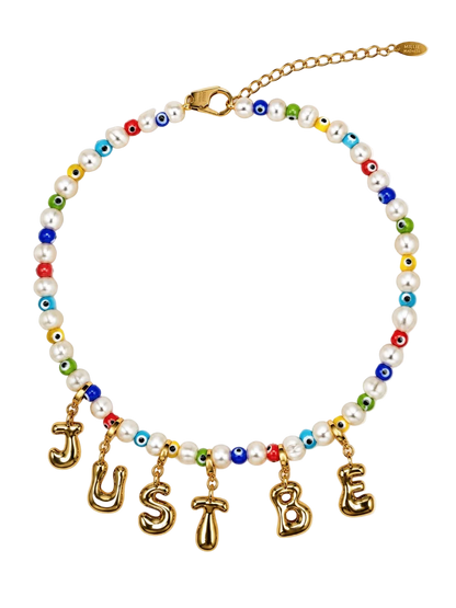A beaded MILLIE MADNESS necklace with white pearl-like beads, colorful evil eye accents, and gold letter charms spelling 'JUST BE' on a white background.