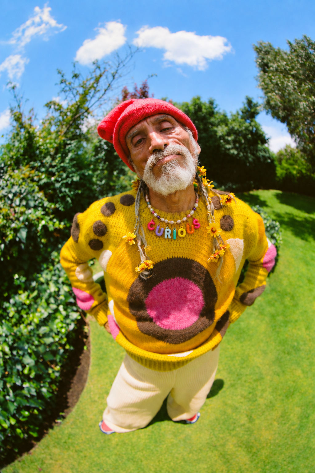 A man outdoors on a grassy lawn under a blue sky, wearing a bright yellow novelty sweater with a pink and brown circular design, a red knit beanie, and multiple colorful necklaces.