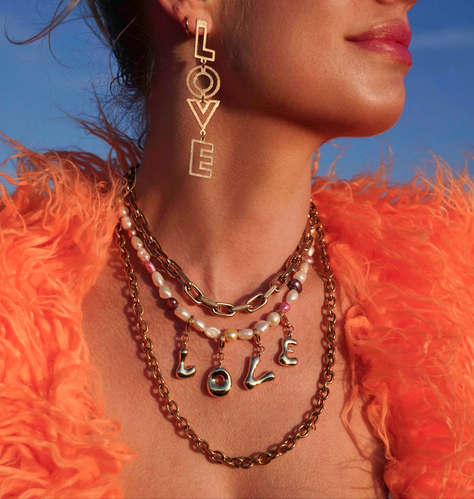 Woman wearing gold necklaces and earrings with 'LOVE' inscription against a blue sky.