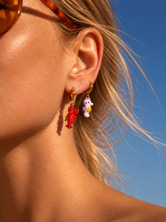 A close-up of a woman's ear wearing two dangling millie madness earrings: a red lobster charm and a small white seahorse charm with yellow details, against a bright blue sky.