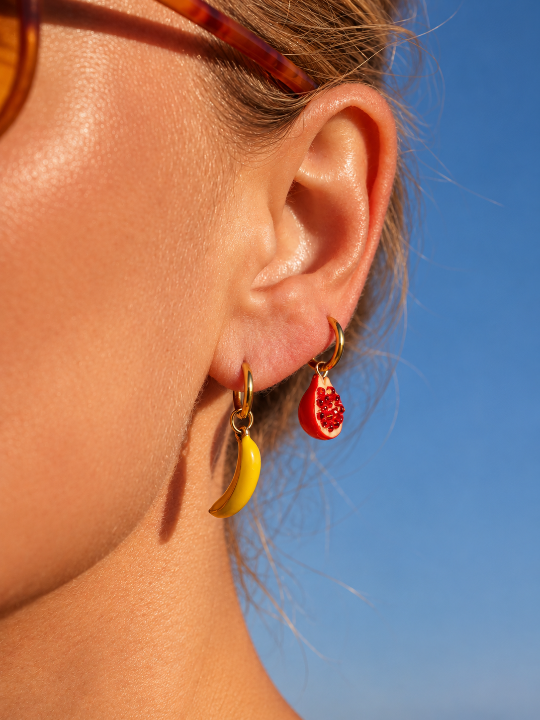 A close-up of a person's ear wearing two small hoop millie madness earrings: one yellow banana-shaped earring and one red pomegranate-shaped earring, against a clear blue sky.