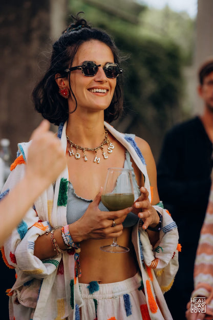 A woman outdoors wearing a colorful bohemian-style embroidered cover-up jacket over a light blue bikini top and white bottoms, holding a glass of green drink. She is wearing dark sunglasses and layered gold millie madness necklaces.