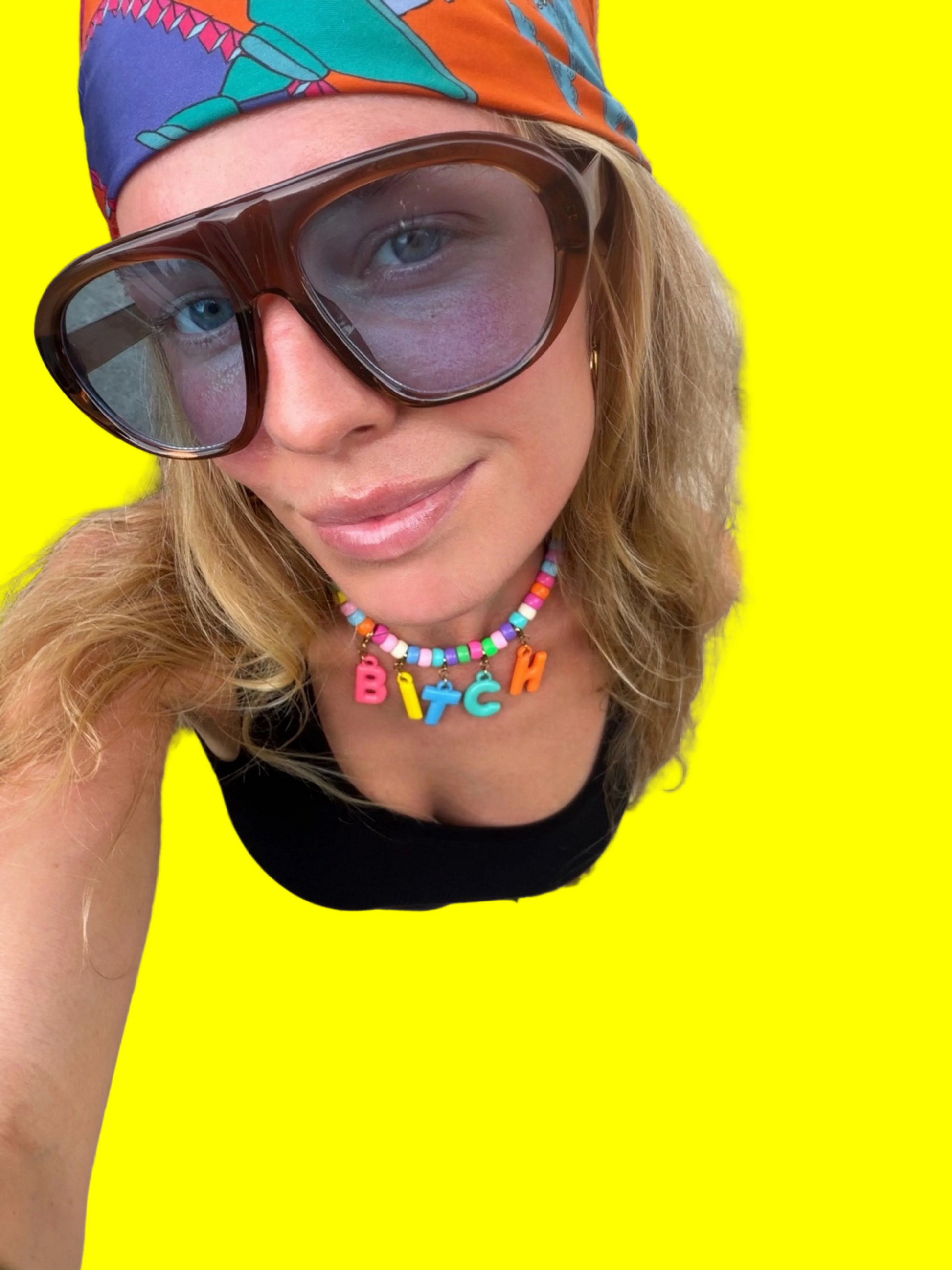 A close-up selfie of a woman wearing a colorful beaded necklace with the word 'BITCH' in letter charms, a black top, and a vibrant patterned headwrap. She is looking down at the camera on a concrete sidewalk.