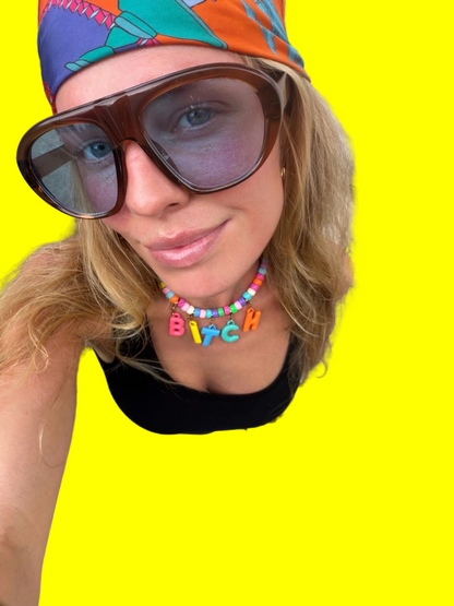 A close-up selfie of a woman wearing a colorful beaded necklace with the word 'BITCH' in letter charms, a black top, and a vibrant patterned headwrap. She is looking down at the camera on a concrete sidewalk.