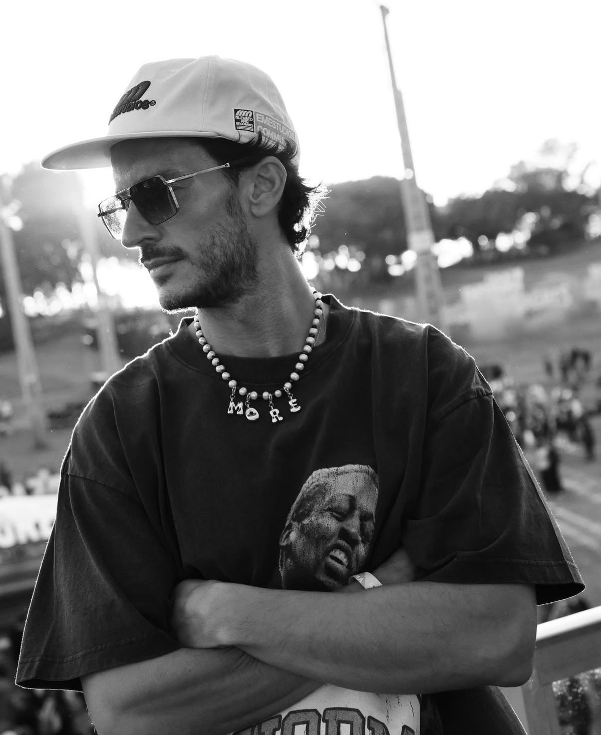 A man wearing a white baseball cap, dark sunglasses, a beaded MILLIE MADNESS NECKLACE with gold letters, and a dark graphic t-shirt, standing with arms crossed in an outdoor stadium setting.
