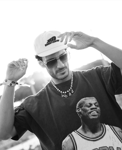 A black and white lifestyle photo of a man wearing a white The North Face cap, sunglasses, a beaded MILLIE MADNESS necklace, and a dark graphic t-shirt featuring a basketball player.