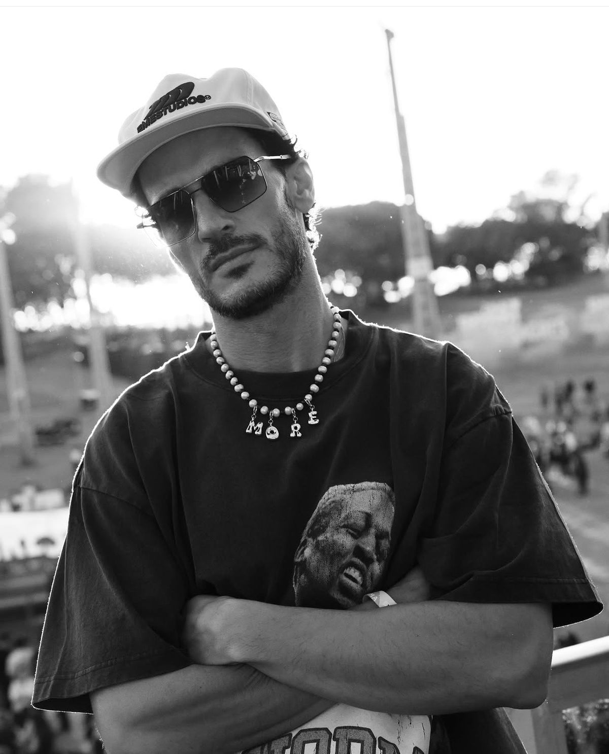 A black and white portrait of a man wearing a baseball cap, sunglasses, a beaded necklace with 'MORE' lettering, and a graphic t-shirt, standing with arms crossed in an outdoor stadium setting.