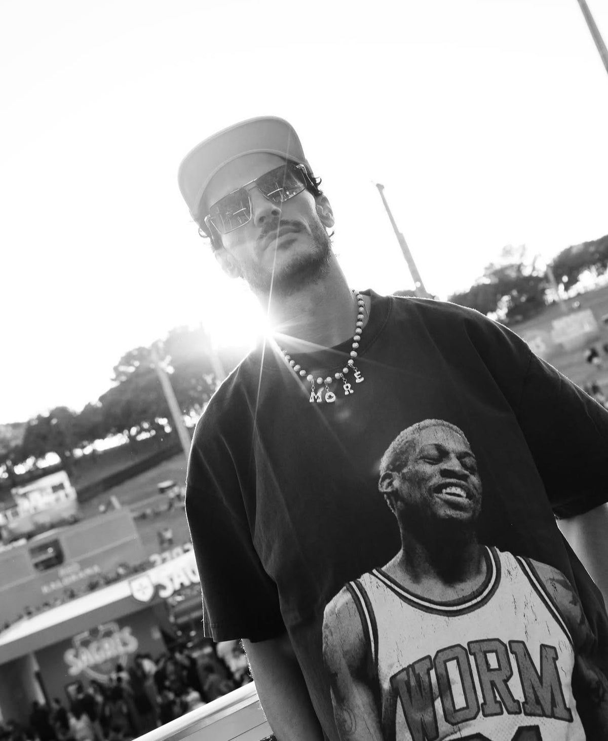 A black and white lifestyle photograph of a man wearing a black t-shirt with a basketball player graphic, a beaded  MILLIE MADNESS necklace, and a cap, set against a bright outdoor stadium background.