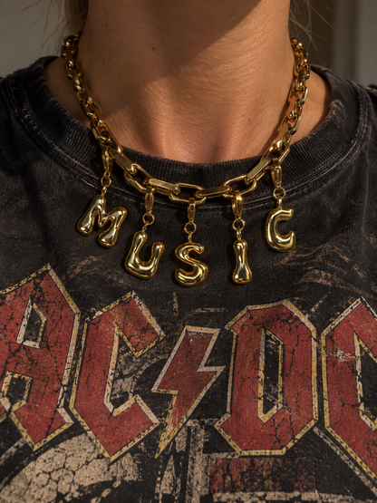 A close-up of a person wearing a chunky gold millie madness chain necklace with dangling letters spelling 'MUSIC' over a black AC/DC graphic t-shirt.