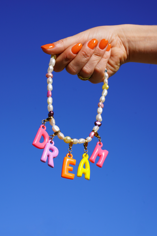 Hand holding a beaded bracelet with colorful letters spelling 'DREAM' against a blue sky.