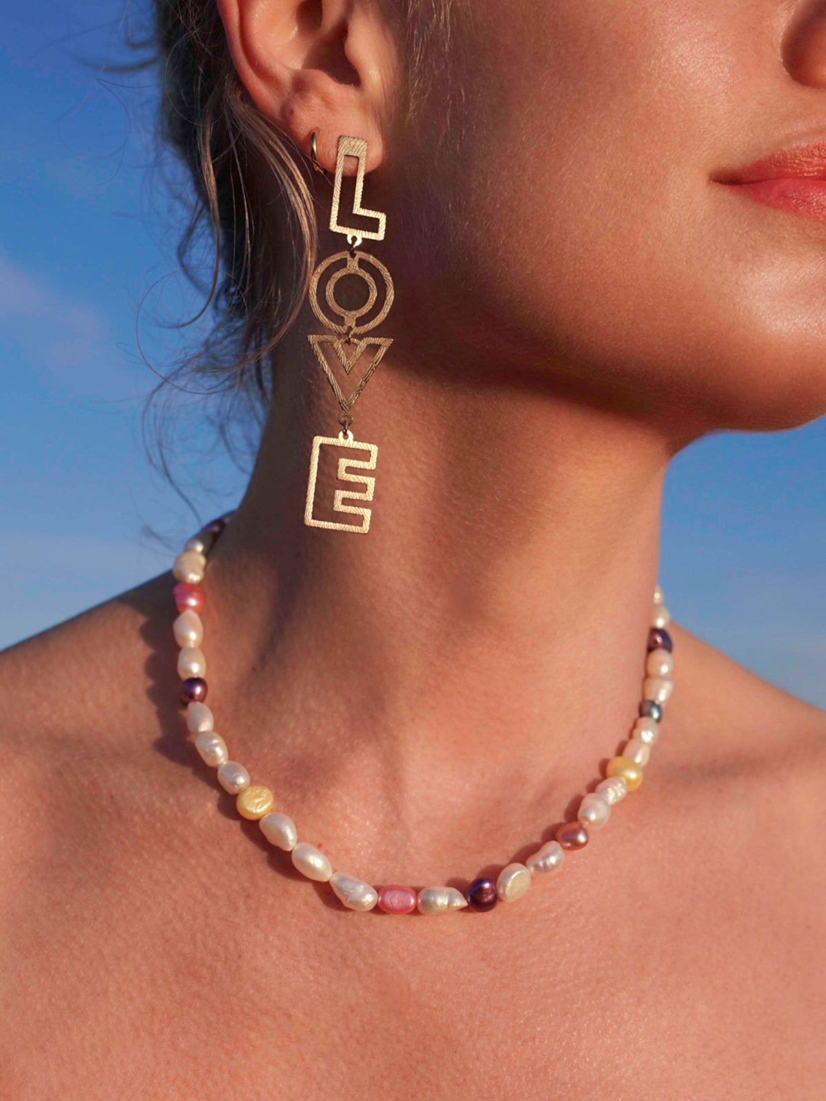 Close-up of a model wearing gold 'LOVE' earrings and a MILLIE MADNESS pearl necklace against a blue sky.