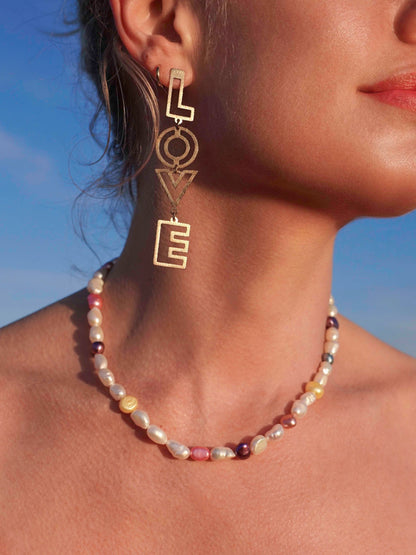 Close-up of a model wearing gold 'LOVE' earrings and a MILLIE MADNESS pearl necklace against a blue sky.
