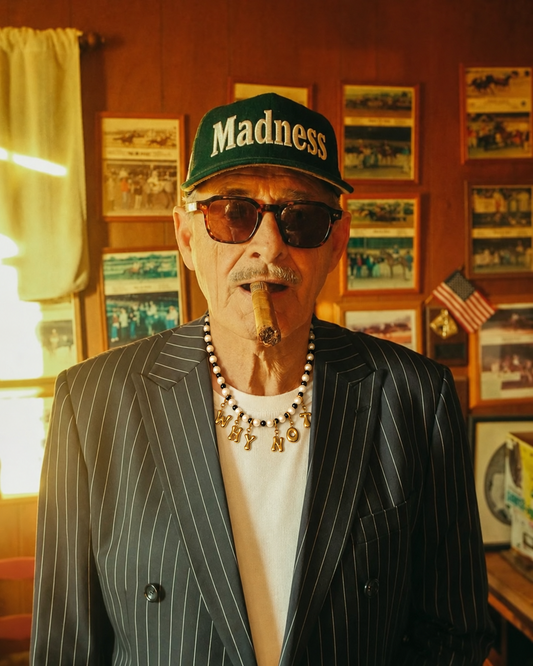 A man wearing a green 'Madness' baseball cap, dark sunglasses, a pinstripe blazer over a white shirt, and a beaded Millie Madness necklace with a cigar in his mouth, standing in a warmly lit room with framed pictures on the wall.