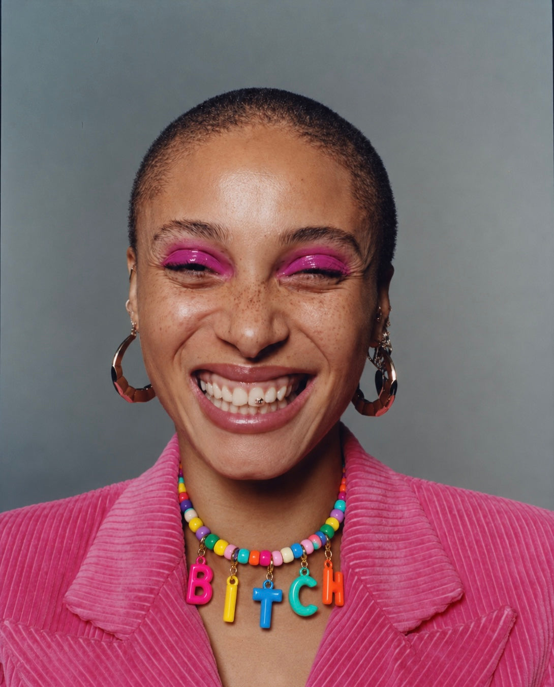 A close-up portrait of a smiling person with a shaved head, wearing a bright pink corduroy jacket, a colorful beaded necklace spelling 'BITCH', and large hoop earrings against a plain gray background.