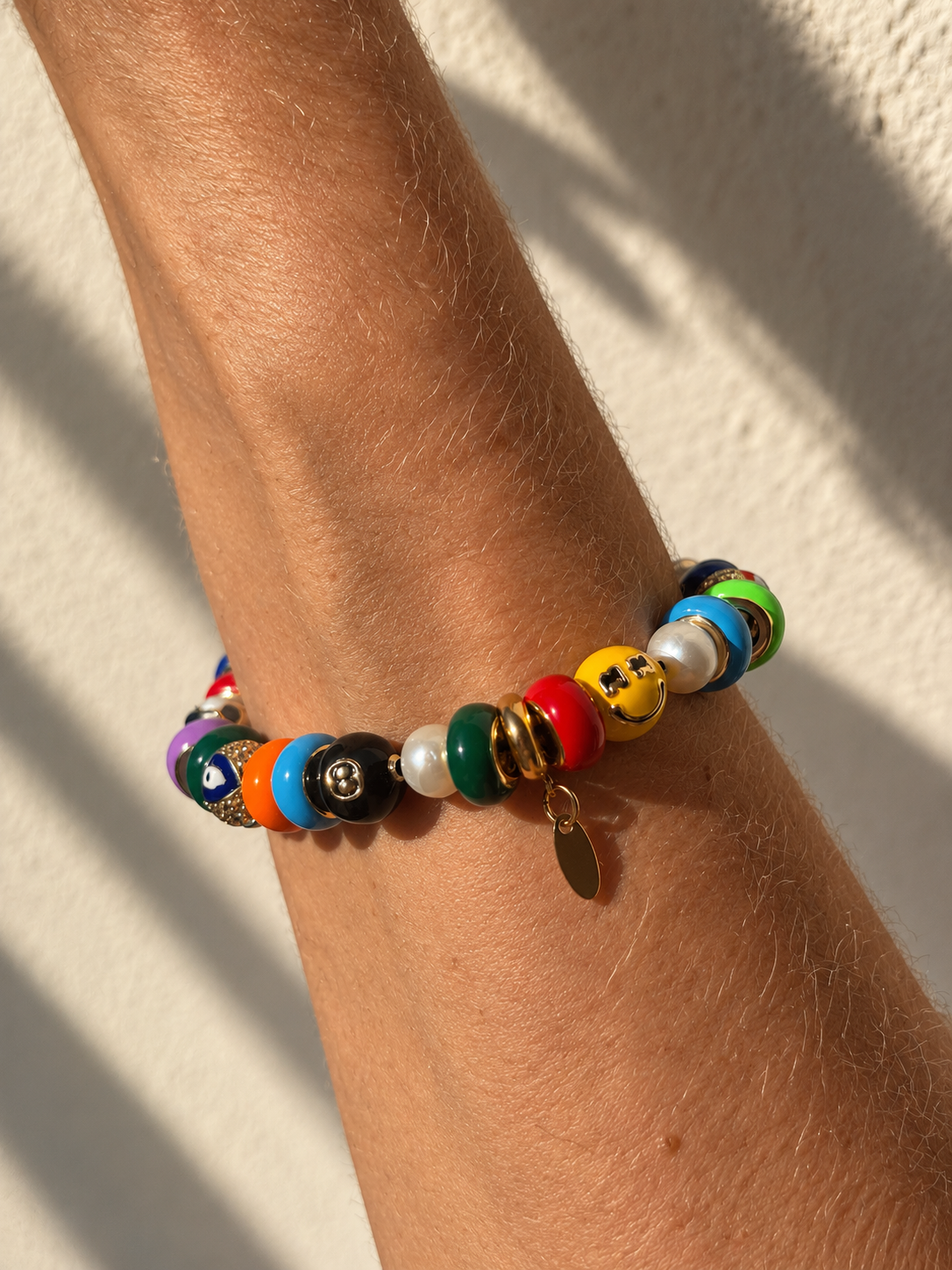 A colorful beaded millie madness bracelet with a small gold charm, shown worn on a person's wrist against a textured beige wall with soft shadows.