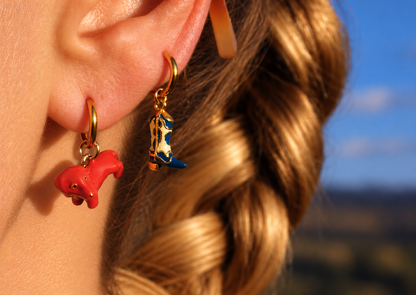 Close-up of a person's ear wearing two dangling earrings: a small red animal charm on a gold hoop and a cowboy boot charm with gold, white, and blue detailing on a gold hook. millie madness