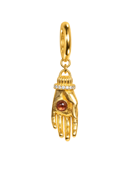 A gold millie madness earring shaped like a hand, featuring a small round amber-red gemstone on the back of the hand and a row of tiny clear stones around the wrist, suspended from a gold ring on a plain white background.