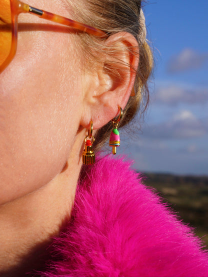 Close-up of a person wearing earrings with a lipstick charm and ice cream charm wearing sunglasses and a pink fur coat with a blurred natural background