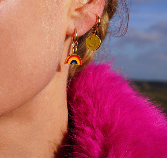 Close-up of an ear with a rainbow earring and a smiley earring against a blurred background