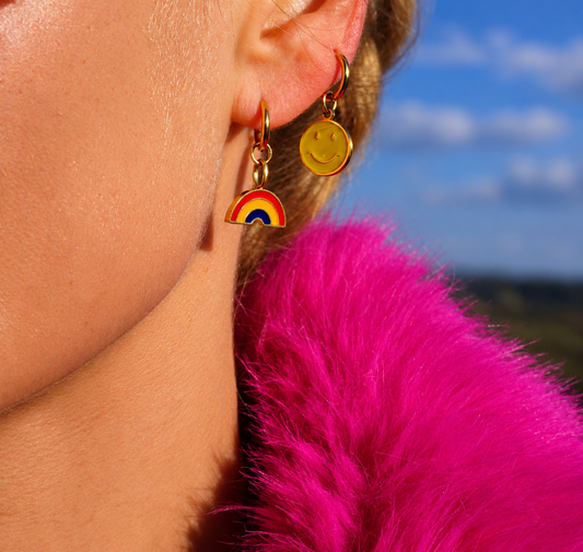 A close-up of a person's ear wearing two gold-tone earrings: a small smiley face charm and a rainbow charm, with a bright pink fuzzy garment in the foreground and a blue sky background.