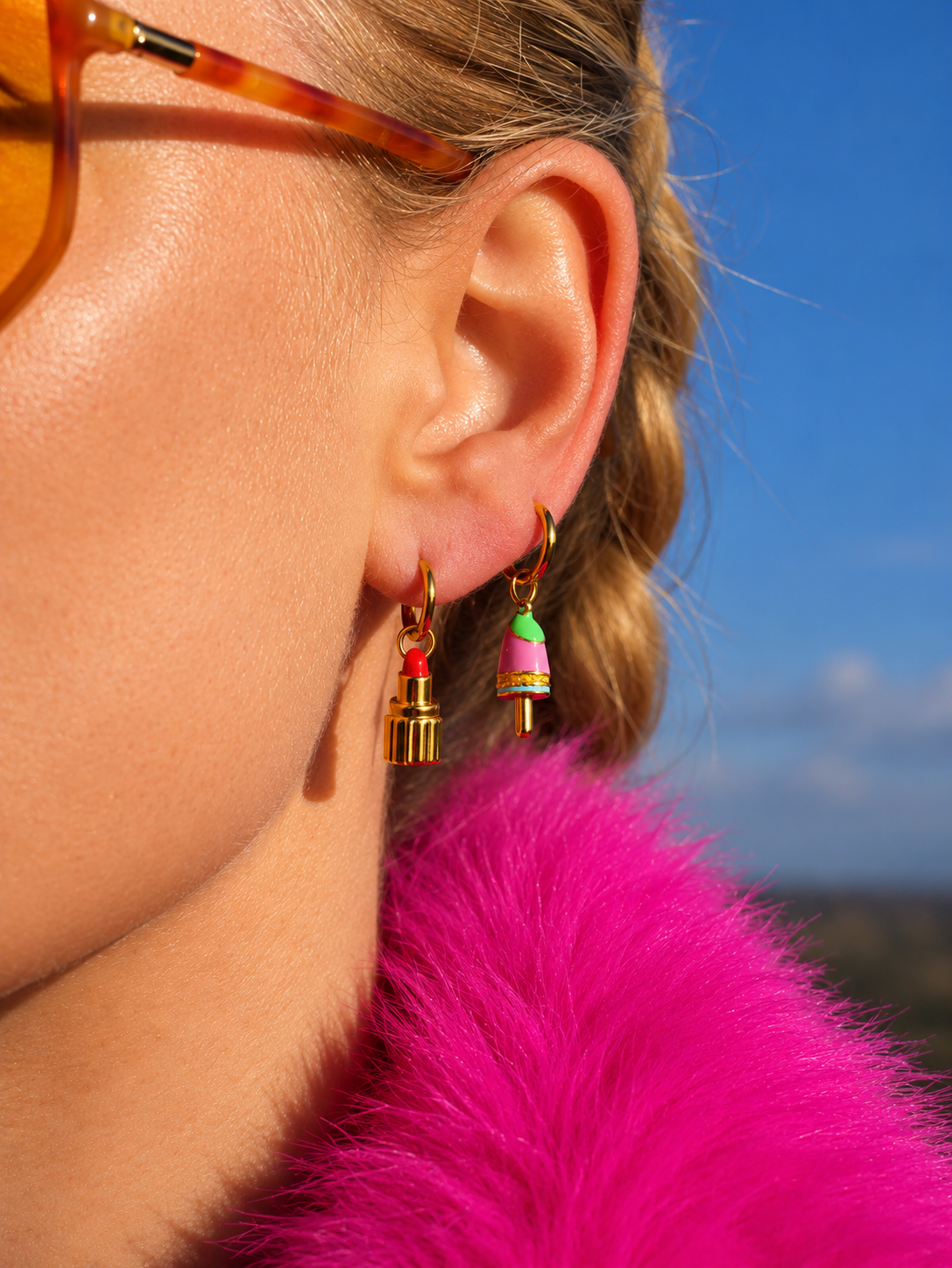 A close-up side profile of a woman wearing colorful bell-shaped millie madness earrings with gold-toned hooks, a bright pink faux fur collar, and amber-tinted sunglasses against a blue sky background.