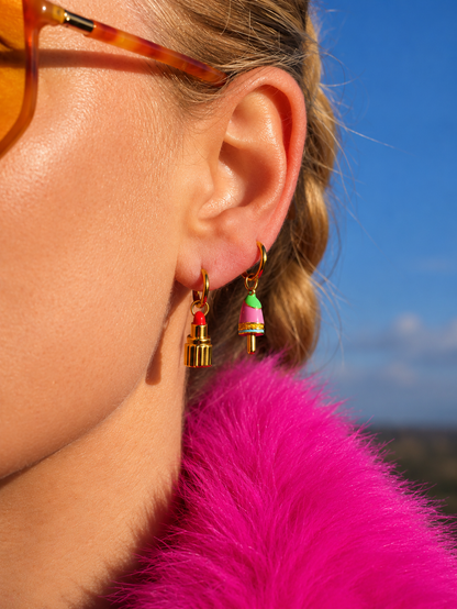 A close-up side profile of a woman wearing colorful bell-shaped millie madness earrings with gold-toned hooks, a bright pink faux fur collar, and amber-tinted sunglasses against a blue sky background.