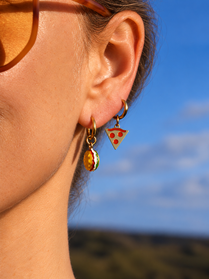 Close-up of a person's ear wearing two gold-tone earrings: a small triangular pizza slice charm with red and yellow detailing, and a dangling round charm with red, yellow, and green pattern. Sunglasses are partially visible, with a blue sky and blurred outdoor background.
