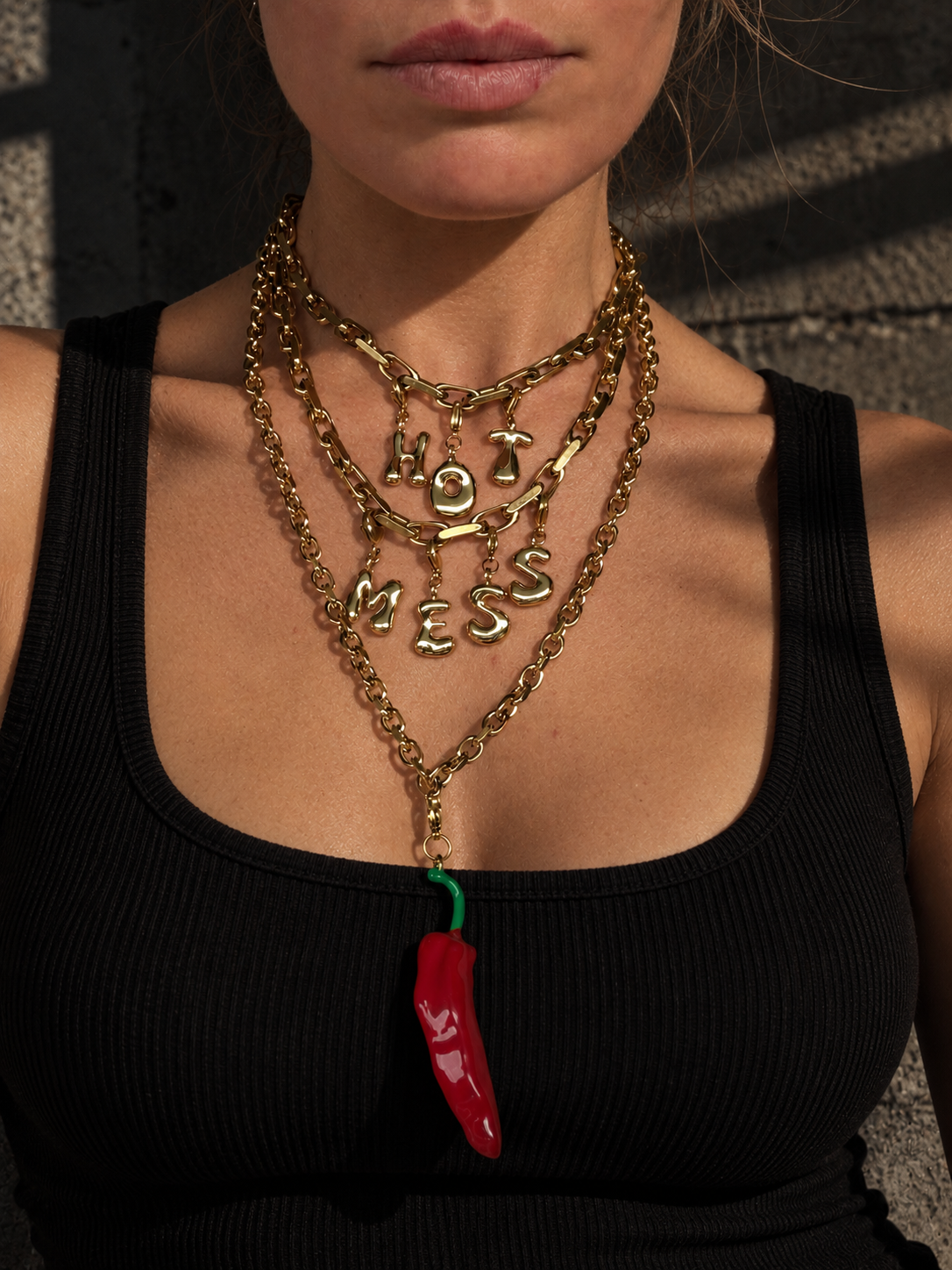A close-up of a model wearing a layered gold necklace set by millie madness. The set includes a 'HOT MESS' letter necklace, a chunky chain necklace, and a longer chain with a dangling red chili pepper charm.