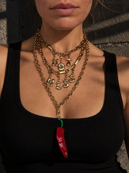 A close-up of a model wearing a layered gold necklace set by millie madness. The set includes a 'HOT MESS' letter necklace, a chunky chain necklace, and a longer chain with a dangling red chili pepper charm.