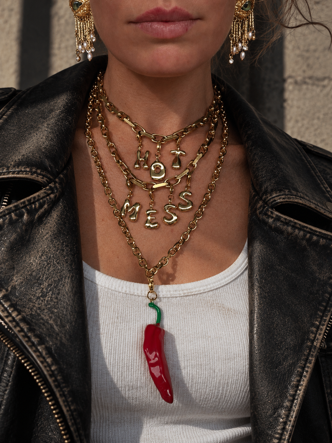 A close-up of a person wearing layered gold-tone millie madness necklaces, including one with the words 'HOT' and 'MESS' in dangling letters, plus a red chili pepper pendant, styled with a black leather jacket and white top.