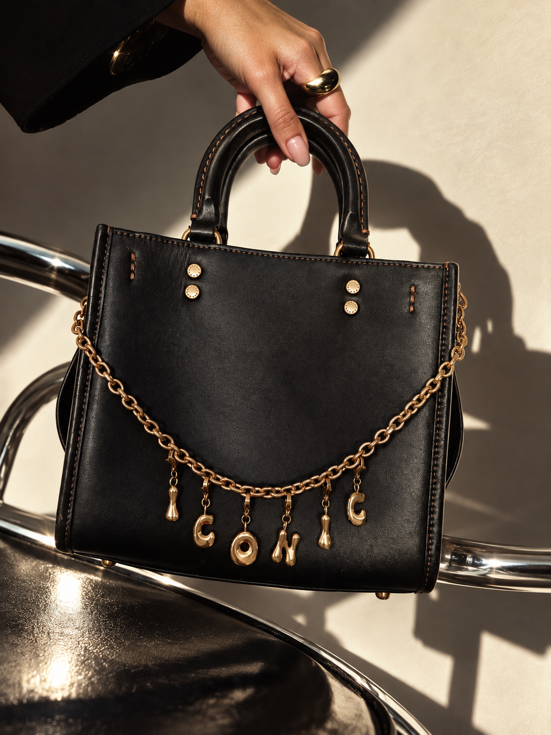 A black leather handbag with gold-tone hardware and a dangling millie madness chain spelling 'ICONIC', held by a person's hand in a styled studio setting with strong sunlight and shadows.