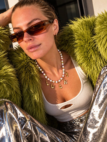 A woman wearing a white ribbed crop top, a green faux fur jacket, and silver metallic pants, accessorized with a millie madness pearl necklace featuring gold initial letters and brown-tinted sunglasses.