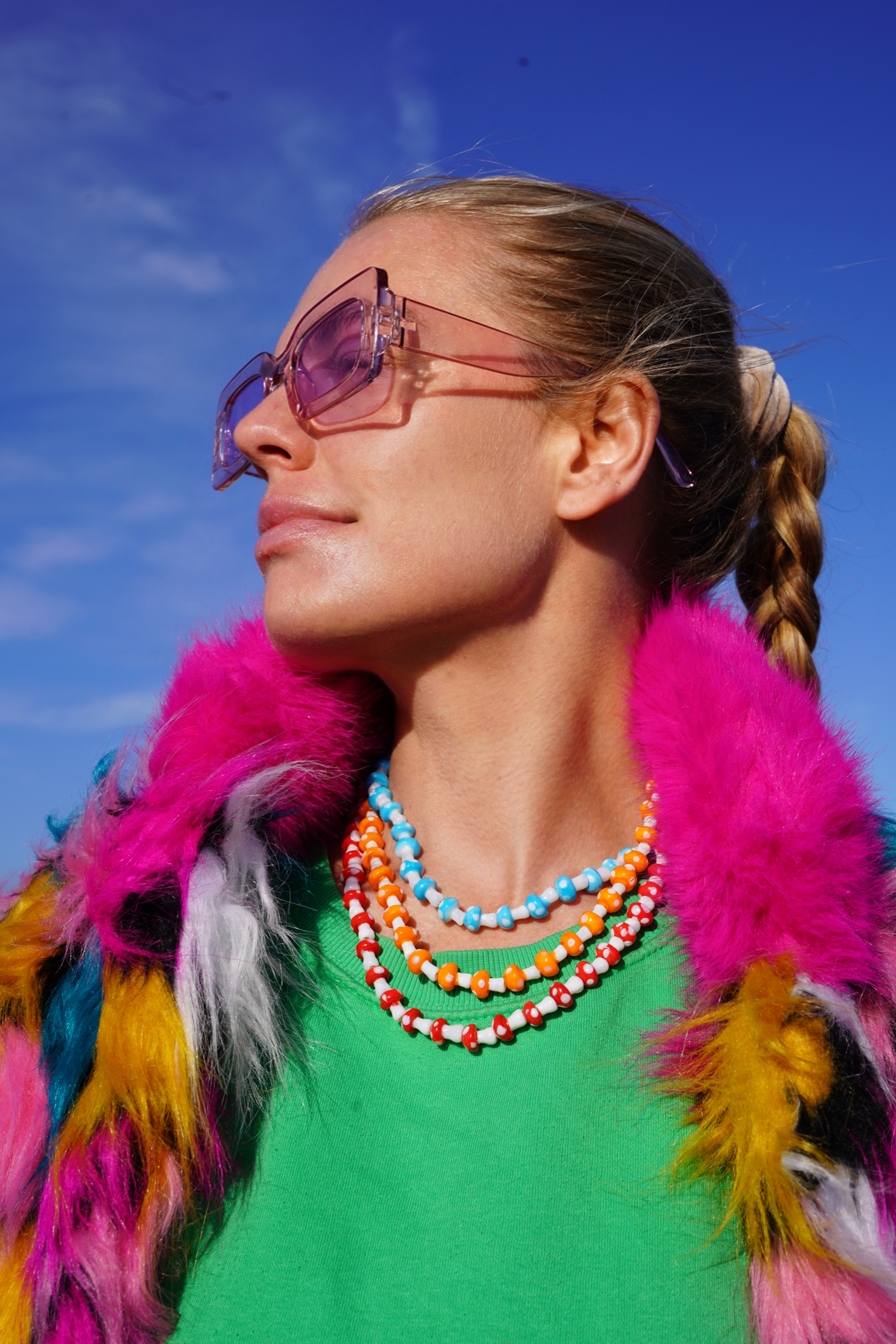 Person wearing colorful sunglasses, pink feathered garment, and multicolored necklace against a blue sky.