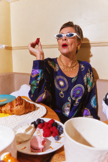 A woman wearing white cat-eye sunglasses and a navy blue patterned blouse and a queen necklace gold chain millie madness necklace sits at a breakfast table, holding a strawberry. Plates with croissants, fruit, and pastries are in the foreground.